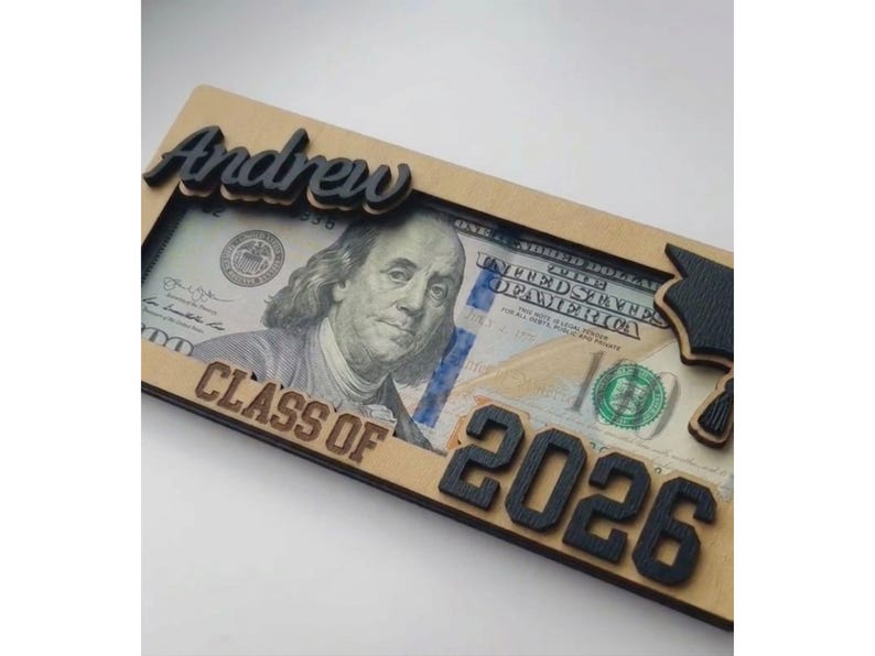 Close-up angled view of a personalized laser-engraved wooden graduation money holder reading "Andrew – Class of 2026," with a $100 bill tucked inside the open slot, showing the raised black script name, bold engraved year numbers, laser-cut graduation cap detail on the right corner, and natural birch wood grain texture against a clean white background.