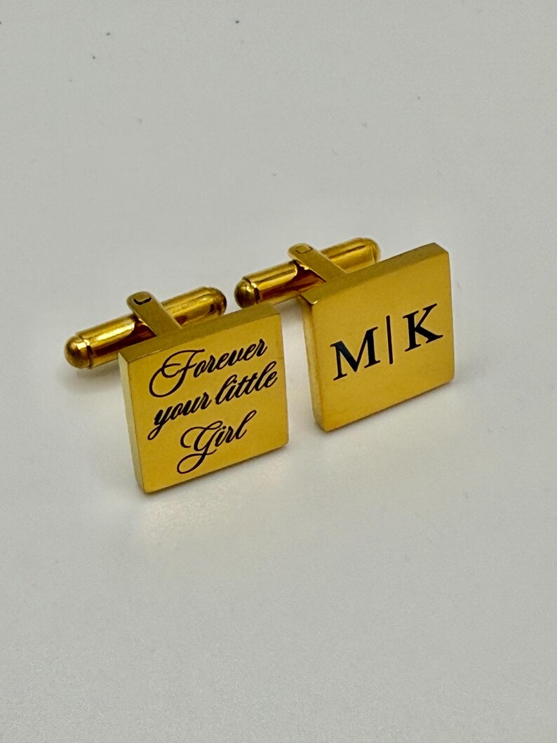 Puede incluir: Gemelos cuadrados dorados. Un gemelo presenta el texto "Forever your little Girl" en escritura cursiva. El otro gemelo muestra las iniciales "M|K" en negro. Los gemelos tienen un poste rectangular y una palanca redondeada.