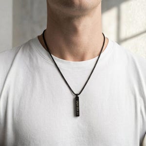 K&ouml;nnte beinhalten: Eine schwarze Halskette mit einem rechteckigen Anh&auml;nger, auf dem die r&ouml;mischen Ziffern VI VIII MMI eingraviert sind. Der Anh&auml;nger h&auml;ngt an einer passenden schwarzen Kette. Die Person tr&auml;gt ein wei&szlig;es T-Shirt.
