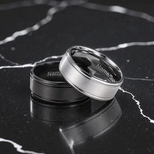 Two men’s titanium wedding bands displayed on a black marble surface—one silver with a brushed center finish and polished edges, and one black with a brushed finish. Both rings feature customizable inner engravings.