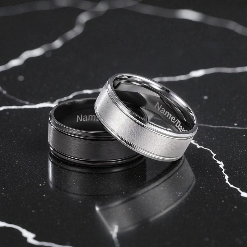 Two men’s titanium wedding bands displayed on a black marble surface—one silver with a brushed center finish and polished edges, and one black with a brushed finish. Both rings feature customizable inner engravings.