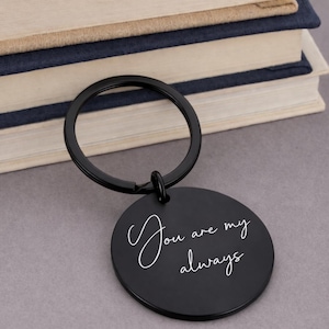 Black round keychain with the words "You are my always" in white script. The keychain is attached to a black ring. The background includes a stack of books.