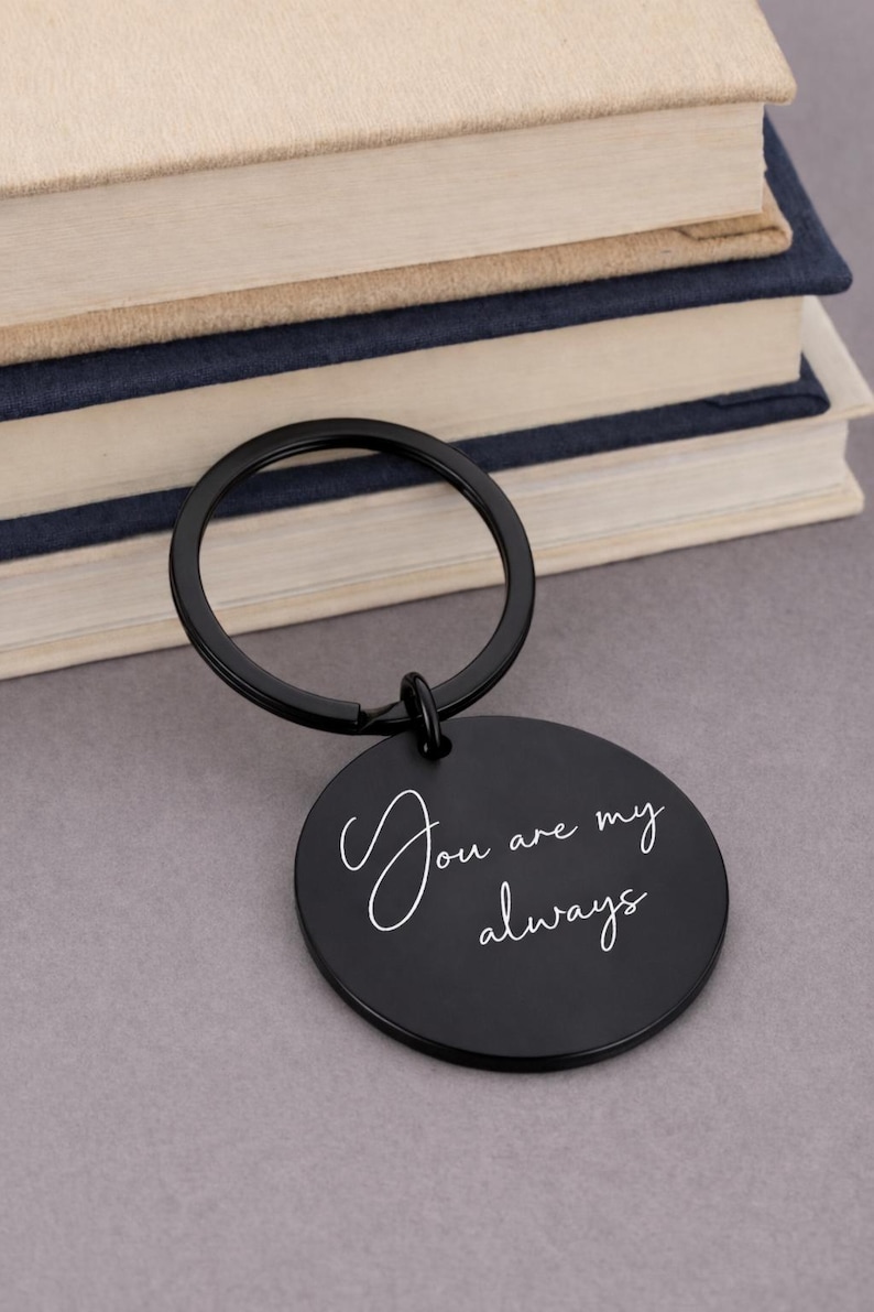 Black round keychain with the words "You are my always" in white script. The keychain is attached to a black ring. The background includes a stack of books.