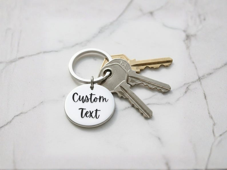 May include: A silver keychain with a round pendant that reads "Custom Text" in a black, cursive font. The keychain is attached to a silver ring with three keys. The background is a white marble surface.