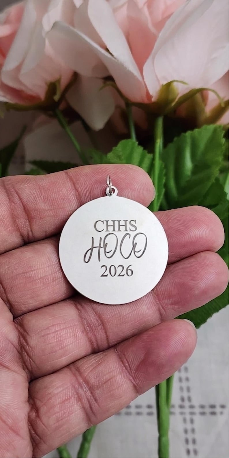 K&ouml;nnte beinhalten: Ein runder Anh&auml;nger aus silberfarbenem Metall mit einer Gravur, die den Text CHHS, HOCO und 2026 zeigt. Der Anh&auml;nger verf&uuml;gt &uuml;ber eine kleine Metall&ouml;se an der Oberseite zur Befestigung und hat eine polierte Oberfl&auml;che mit dunkler Schrift.