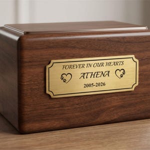 K&ouml;nnte beinhalten: Rechteckige Holzurne mit polierter Oberfl&auml;che. Auf einer goldfarbenen Plakette steht "FOREVER IN OUR HEARTS" &uuml;ber dem Namen "ATHENA" und den Daten "2005-2026", mit Herz- und Pfotenabdrucksymbolen.