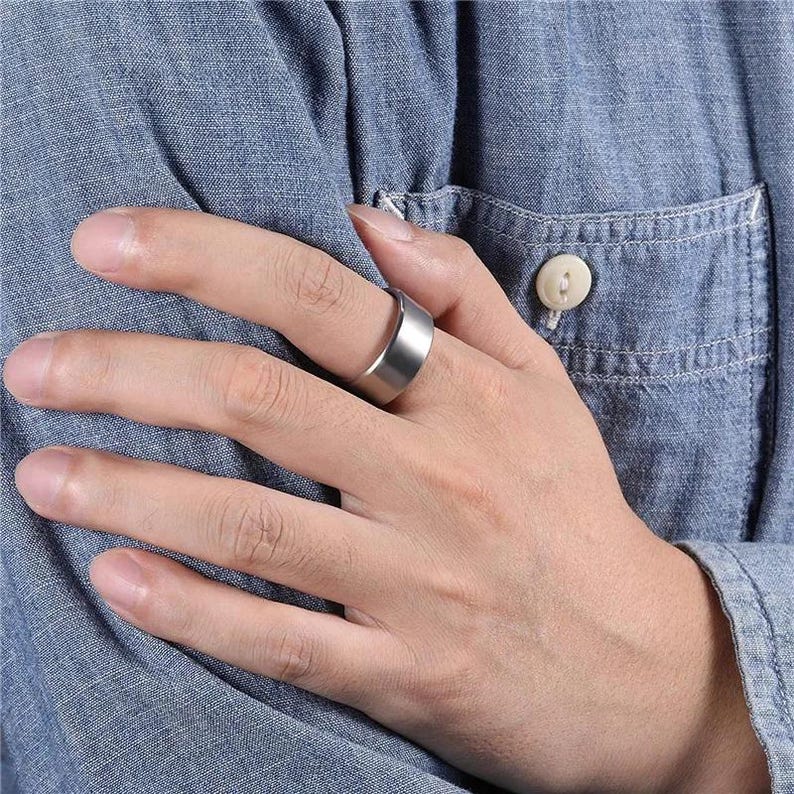 A gold-colored ring with a flat top and beveled edges. The ring is worn on a finger, showcasing its simple, modern design. The ring's surface has a matte finish, reflecting light subtly.