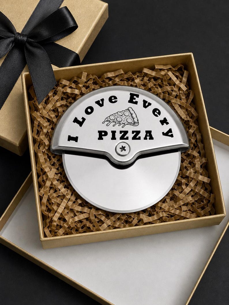 Stainless steel pizza cutter engraved with “I Love Every Pizza” and a pizza slice illustration, neatly placed in a gold gift box with brown crinkle paper filler and a black ribbon lid.