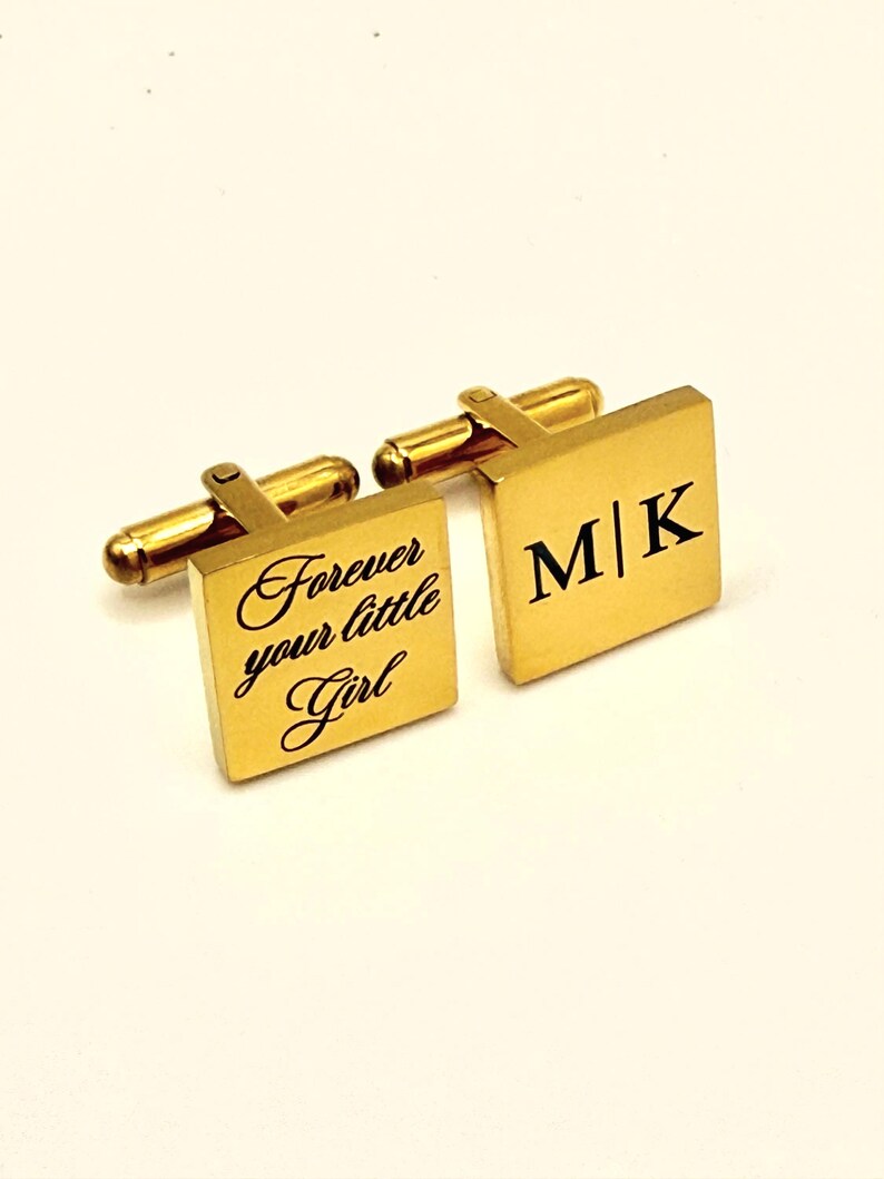 Gold-toned square cufflinks. One cufflink features the text "Forever your little Girl" in cursive script. The other cufflink displays the initials "M|K" in a bold, black font. The cufflinks have a classic, elegant design.