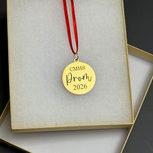 May include: A gold-colored round ornament with the text "CMHS Prom 2026" in a decorative font, suspended by a red ribbon. The ornament is presented inside a beige gift box with a white, textured interior.