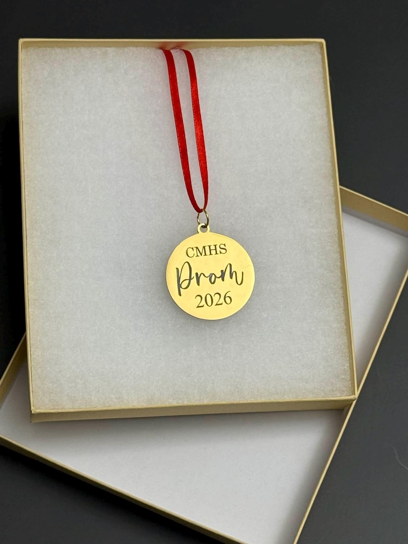 May include: A gold-colored round ornament with the text "CMHS Prom 2026" in a decorative font, suspended by a red ribbon. The ornament is presented inside a beige gift box with a white, textured interior.