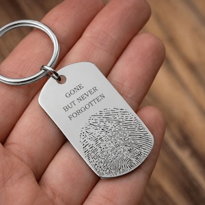 Close-up of a silver stainless steel memorial keychain resting in an open hand. The rectangular dog-tag-style keychain features the engraved message “Gone But Never Forgotten” above a detailed fingerprint engraving. The keyring is attached through a small hole at the top corner, and the background shows a softly blurred wooden surface, creating a warm and sentimental presentation.