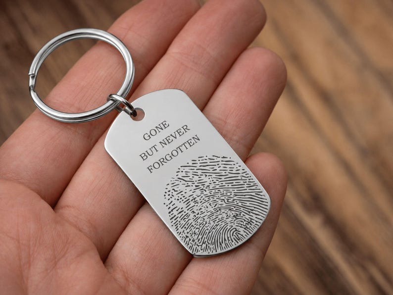 Close-up of a silver stainless steel memorial keychain resting in an open hand. The rectangular dog-tag-style keychain features the engraved message “Gone But Never Forgotten” above a detailed fingerprint engraving. The keyring is attached through a small hole at the top corner, and the background shows a softly blurred wooden surface, creating a warm and sentimental presentation.