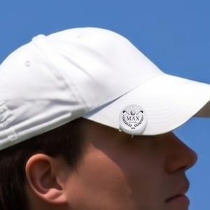 Close-up profile view of a male golfer wearing a white hat with a personalized magnetic golf ball marker clipped to the brim. The custom silver marker features an engraved laurel wreath logo, golf clubs, and the text "MAX BY PAR 2026" against a clear blue sky.