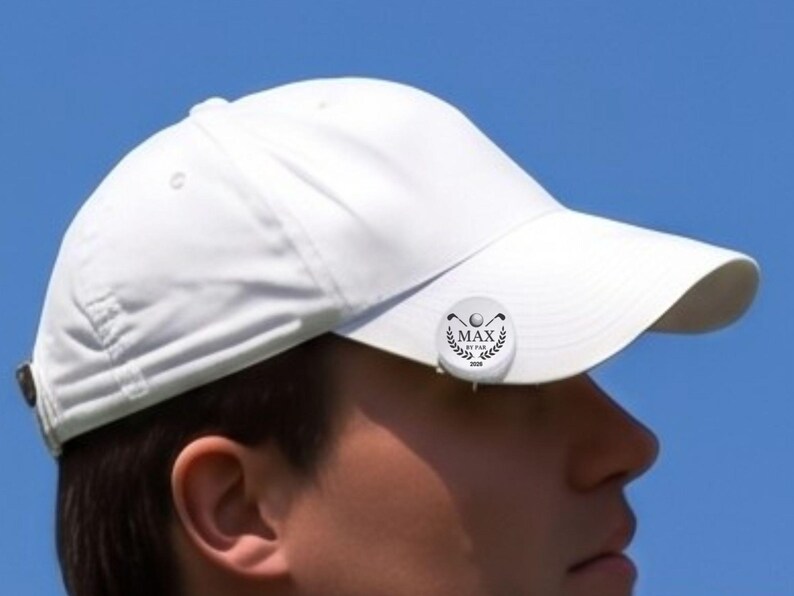 Close-up profile view of a male golfer wearing a white hat with a personalized magnetic golf ball marker clipped to the brim. The custom silver marker features an engraved laurel wreath logo, golf clubs, and the text "MAX BY PAR 2026" against a clear blue sky.