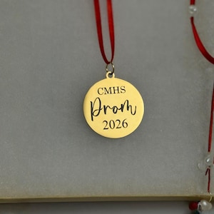 May include: A gold-colored round pendant with the word "Prom" in cursive and the year "2026" engraved on it. The pendant is attached to a red ribbon. The text "CMHS" is above the word "Prom".