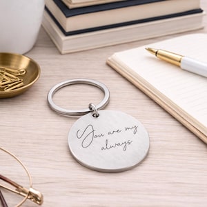 Round stainless steel engraved keychain resting on a light wood desk, featuring the handwritten message “You are my always.” The keychain is styled in a cozy workspace setting with stacked books, a notebook and pen, a small potted plant, paper clips, and eyeglasses in the background. Soft natural lighting and a neutral color palette create an elegant, sentimental gift presentation.