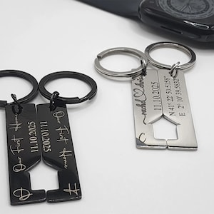 Two pairs of metal keychains displayed on a white surface, one black and one silver. Each pair has interlocking rectangular pieces with cutout shapes forming a house when joined. The black keychains are engraved with “Our First Home” and the date “11.10.2025,” while the silver ones are engraved with coordinates, the same date, and a heart design with names. A black smartwatch is visible in the background.