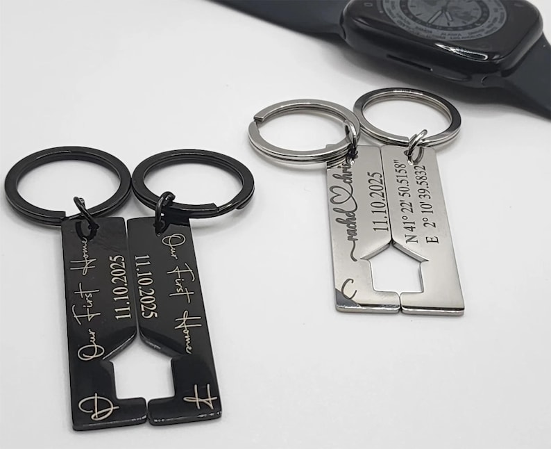 Two pairs of metal keychains displayed on a white surface, one black and one silver. Each pair has interlocking rectangular pieces with cutout shapes forming a house when joined. The black keychains are engraved with “Our First Home” and the date “11.10.2025,” while the silver ones are engraved with coordinates, the same date, and a heart design with names. A black smartwatch is visible in the background.