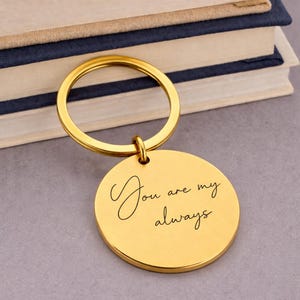 A gold-colored keychain with a round pendant. The pendant has the inscription "You are my always" in elegant script. The keychain is resting on a gray surface with a stack of books in the background.