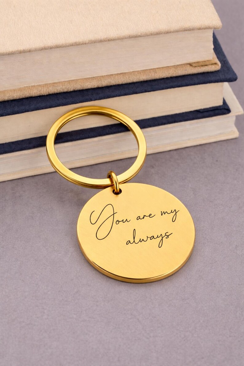 A gold-colored keychain with a round pendant. The pendant has the inscription "You are my always" in elegant script. The keychain is resting on a gray surface with a stack of books in the background.