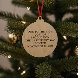 May include: Wooden Christmas ornament with a round shape and a red ribbon for hanging. The ornament has the text "DUE TO THE HIGH COST OF PRODUCTION. THE LAST PENNY WAS MINTED NOVEMBER 12, 2025" in black font. The ornament hangs on a Christmas tree.