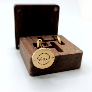 Gold cufflinks with a personalized inscription, presented in a dark wooden box. The cufflinks are round with a gold finish, engraved with names and a date. The box is open, revealing the cufflinks and their storage.