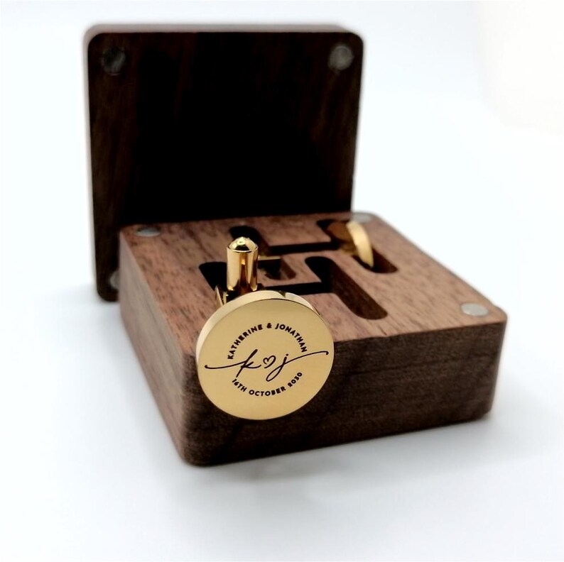Gold cufflinks with a personalized inscription, presented in a dark wooden box. The cufflinks are round with a gold finish, engraved with names and a date. The box is open, revealing the cufflinks and their storage.