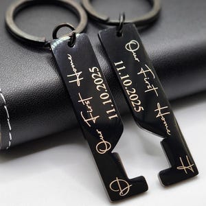 Personalized black metal keychains engraved with “Our First Home” and the date “11.10.2025,” featuring a heart design. Displayed on a leather wraped whiskey flask, these custom keychains make a meaningful housewarming or new home gift for couples.