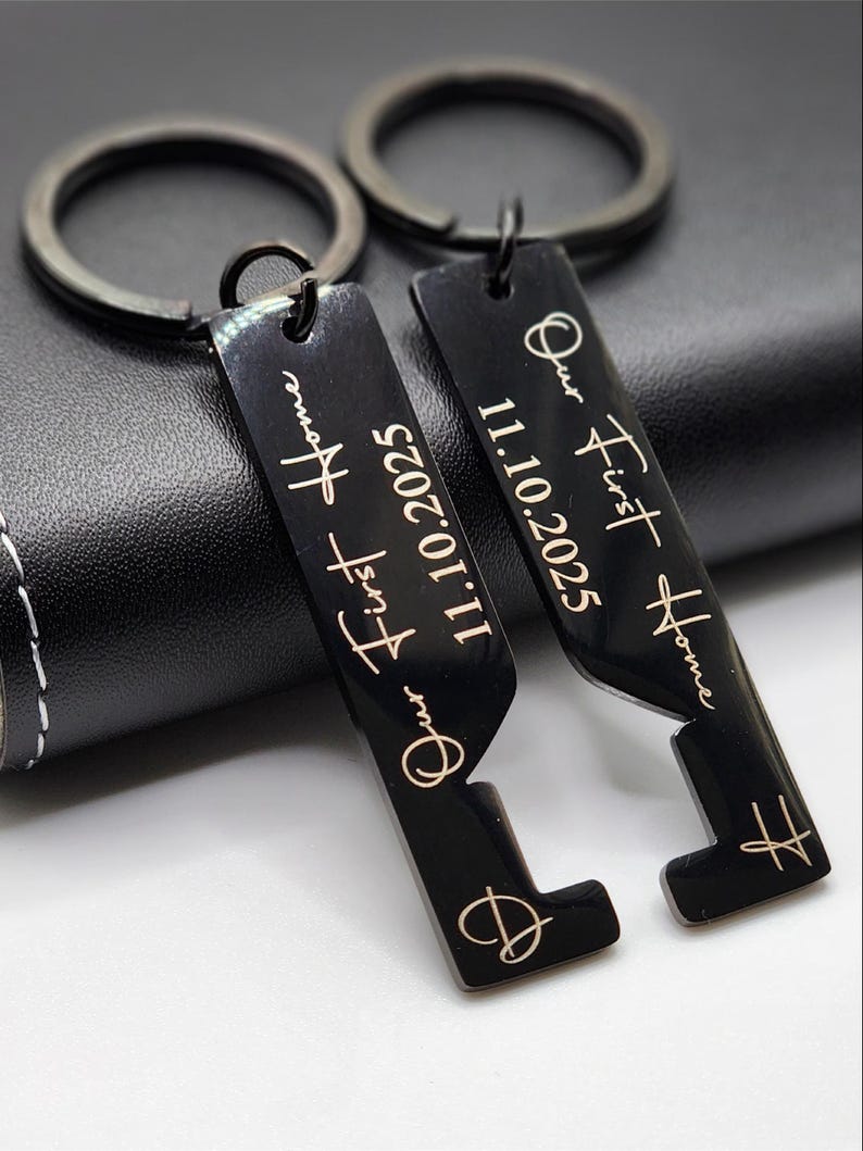 Personalized black metal keychains engraved with “Our First Home” and the date “11.10.2025,” featuring a heart design. Displayed on a leather wraped whiskey flask, these custom keychains make a meaningful housewarming or new home gift for couples.