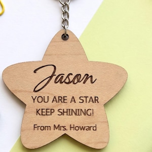 Laser-cut wooden star-shaped keychain engraved with the name "Jason" in script lettering and the message "YOU ARE A STAR KEEP SHINING! From Mrs. Howard." The keychain is attached to a metal chain and displayed on a clean white and pastel green background, photographed in a flat-lay style.