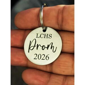 A person's hand is holding a circular stainless steel keychain between their thumb and fingers. The keychain is laser-engraved with black text that reads "LCHS" in a clean serif font at the top, "Prom" in a stylish cursive script in the middle, and the year "2026" in a matching serif font at the bottom.

The metal has a brushed finish, and a sturdy silver split ring is attached through a small hole at the top. The background is softly blurred, keeping the focus entirely on the engraved charm.
