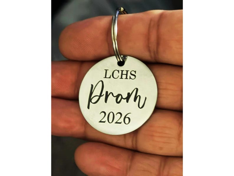 A person's hand is holding a circular stainless steel keychain between their thumb and fingers. The keychain is laser-engraved with black text that reads "LCHS" in a clean serif font at the top, "Prom" in a stylish cursive script in the middle, and the year "2026" in a matching serif font at the bottom.

The metal has a brushed finish, and a sturdy silver split ring is attached through a small hole at the top. The background is softly blurred, keeping the focus entirely on the engraved charm.