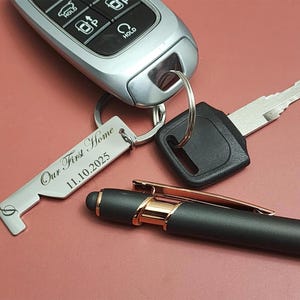 A silver keychain with the text "Our First Home 11.10.2025" is attached to a car key and a car key fob. A black pen with gold accents rests nearby on a maroon surface.