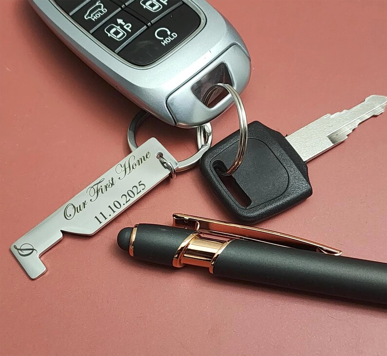 A silver keychain with the text "Our First Home 11.10.2025" is attached to a car key and a car key fob. A black pen with gold accents rests nearby on a maroon surface.
