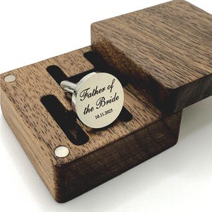 A silver cufflink engraved with "Father of the Bride" and the date 10.11.2025, presented in a dark wooden box. The box has a hinged lid and a custom-fitted interior to hold the cufflink securely. The cufflink is round with a polished finish.