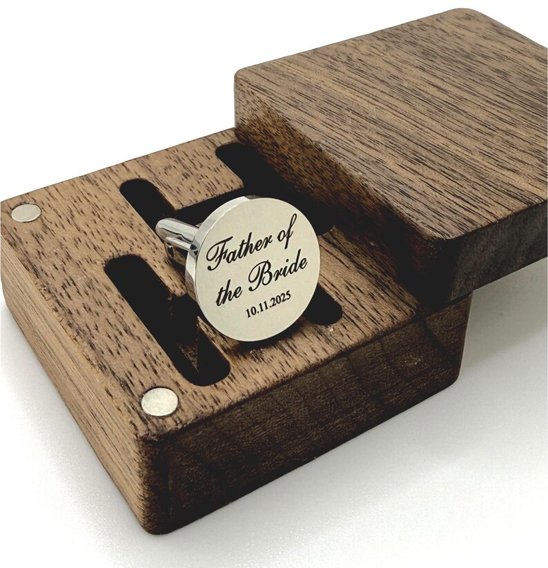 A silver cufflink engraved with "Father of the Bride" and the date 10.11.2025, presented in a dark wooden box. The box has a hinged lid and a custom-fitted interior to hold the cufflink securely. The cufflink is round with a polished finish.