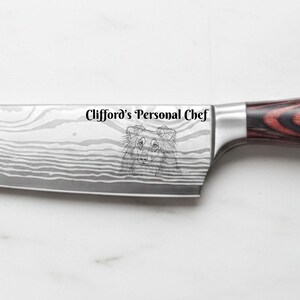 Personalized Damascus-style chef knife with a red and black pakkawood handle, custom laser engraved with the text "Clifford's Personal Chef" and a detailed portrait of a dog on the blade, displayed on a white marble background.