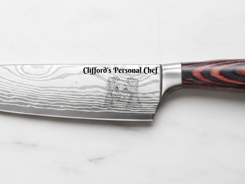 Personalized Damascus-style chef knife with a red and black pakkawood handle, custom laser engraved with the text "Clifford's Personal Chef" and a detailed portrait of a dog on the blade, displayed on a white marble background.