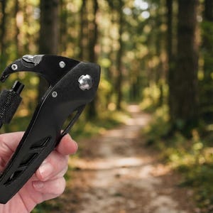 A hand is holding a black carabiner multi-tool, which is made of metal and plastic. The multi-tool has several features, including a bottle opener, a wrench, a Phillips screwdriver, and a flathead screwdriver. The tool is held against a background of a forest trail. The forest is dense with trees and green foliage. The trail is unpaved and looks like a hiking path. The multi-tool is the main focus of the image, and the background is slightly blurred. The lighting is natural and diffuse.