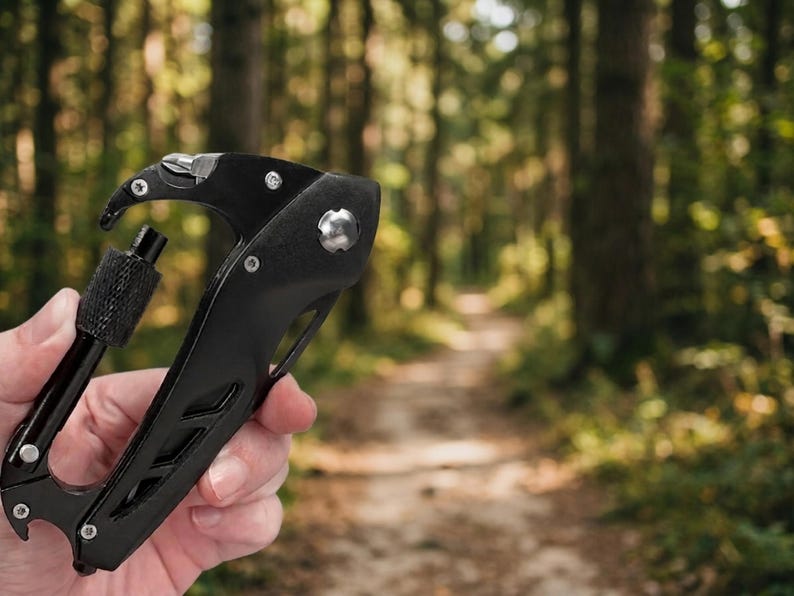 A hand is holding a black carabiner multi-tool, which is made of metal and plastic. The multi-tool has several features, including a bottle opener, a wrench, a Phillips screwdriver, and a flathead screwdriver. The tool is held against a background of a forest trail. The forest is dense with trees and green foliage. The trail is unpaved and looks like a hiking path. The multi-tool is the main focus of the image, and the background is slightly blurred. The lighting is natural and diffuse.