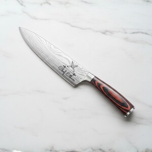 A stainless steel chef's knife with a patterned blade and a red and black wooden handle. The knife has the text "Chef Ethan" etched on the blade. The knife is angled on a white marble surface.