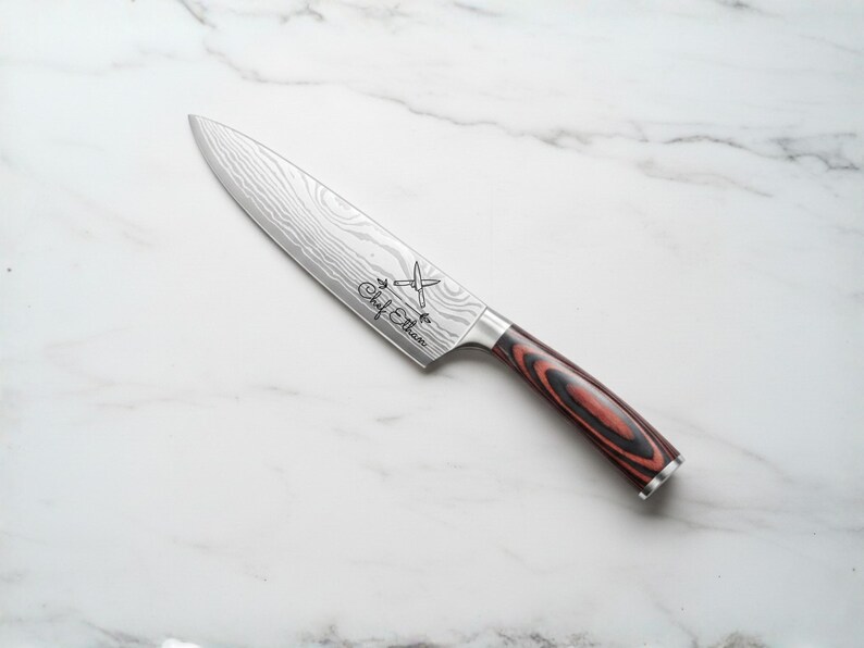 A stainless steel chef's knife with a patterned blade and a red and black wooden handle. The knife has the text "Chef Ethan" etched on the blade. The knife is angled on a white marble surface.