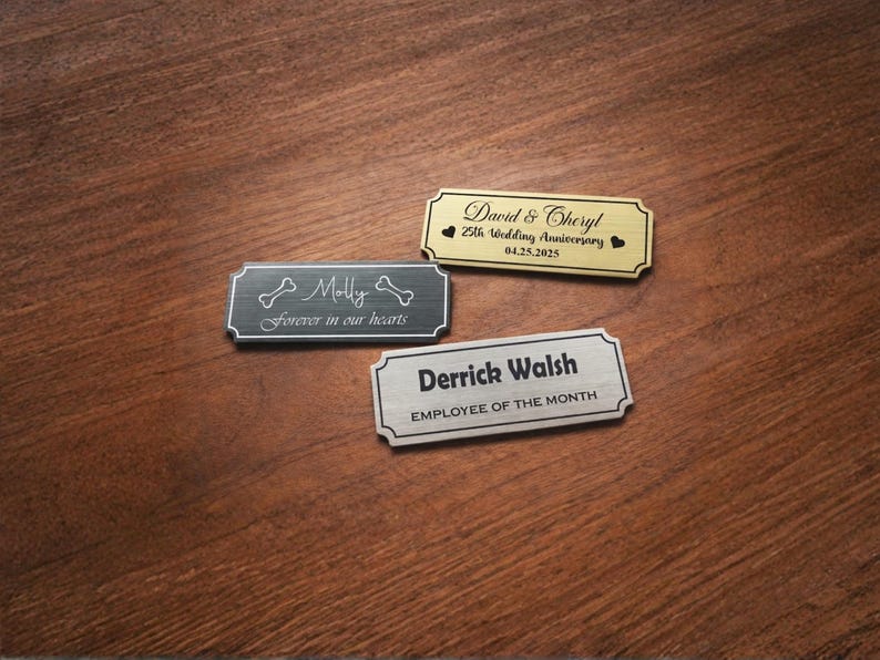 Three engraved metal nameplates displayed on a wooden table: a black pet memorial plaque reading “Molly – Forever in our hearts” with bone icons, a gold “David & Cheryl – 25th Wedding Anniversary – 04.25.2025” plate with heart symbols, and a silver “Derrick Walsh – Employee of the Month” nameplate, all with decorative borders.