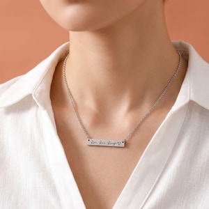 A close-up of a woman wearing a personalized silver horizontal bar necklace. The delicate silver chain holds a sleek, rectangular bar pendant engraved with the words "Live. Love. Laugh." followed by a small heart symbol in a script font. She is wearing a white linen collared shirt against a soft terracotta background.