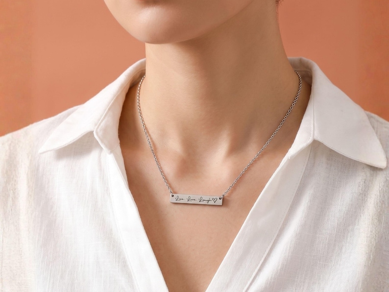 A close-up of a woman wearing a personalized silver horizontal bar necklace. The delicate silver chain holds a sleek, rectangular bar pendant engraved with the words "Live. Love. Laugh." followed by a small heart symbol in a script font. She is wearing a white linen collared shirt against a soft terracotta background.