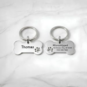 Front and back view of a personalized stainless steel bone-shaped pet ID dog tag with custom laser engraving. The front displays the pet name "Thomas" with a paw print, while the back includes "Microchipped," owner address, and phone number. Durable custom dog name tag with split key ring for pet identification, engraved pet accessory, and thoughtful gift for dog owners.