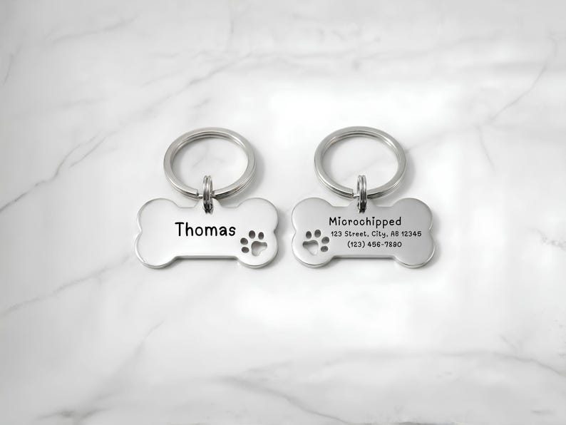 Front and back view of a personalized stainless steel bone-shaped pet ID dog tag with custom laser engraving. The front displays the pet name "Thomas" with a paw print, while the back includes "Microchipped," owner address, and phone number. Durable custom dog name tag with split key ring for pet identification, engraved pet accessory, and thoughtful gift for dog owners.