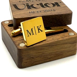 Puede incluir: Un gemelo cuadrado dorado con las iniciales "M|K" se muestra en una caja de madera. La tapa de la caja est&aacute; grabada con "To My Groom" y el nombre "Victor" junto con la fecha "10-11-2025". La caja es de madera oscura.