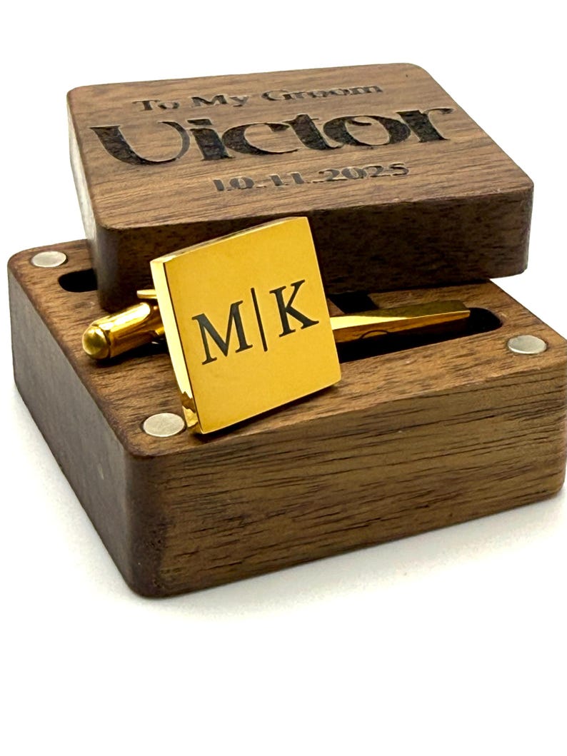 Puede incluir: Un gemelo cuadrado dorado con las iniciales "M|K" se muestra en una caja de madera. La tapa de la caja est&aacute; grabada con "To My Groom" y el nombre "Victor" junto con la fecha "10-11-2025". La caja es de madera oscura.